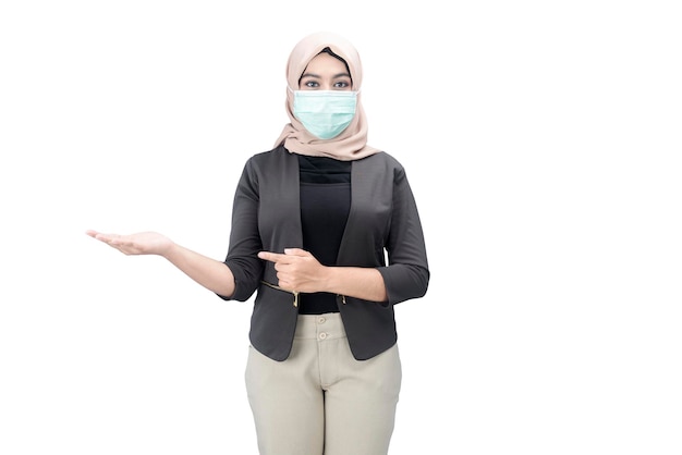 Азиатская мусульманка в платке и в маске для лица стоит с открытыми ладонями, показывая что-то изолированное на белом фоне