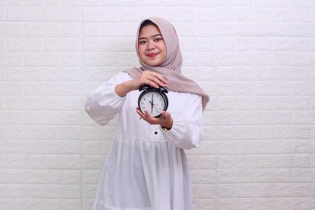Asian Muslim woman in hijab holds an alarm clock in hands showing iftar time 6 pm