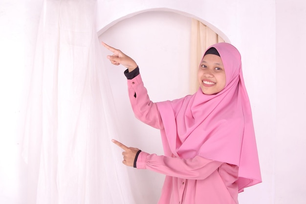 Asian Muslim woman in pink dress pointing sideways to negative advertising space, clipping path.