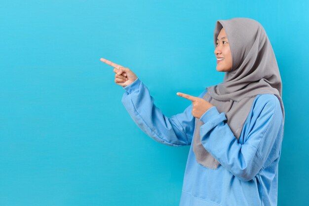 Asian muslim woman points away on copy space with hijab feels optimistic isolated over blue background