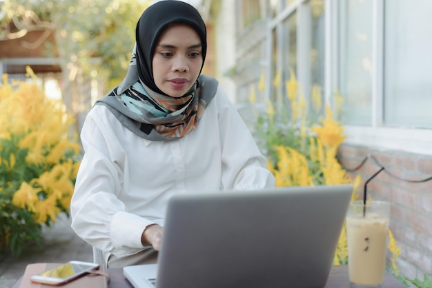 Asian muslim woman sits in a garden with a laptop and a phone on her lap