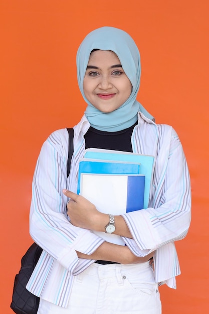 Asian muslim woman standing with backpack holding books isolated on orange background.