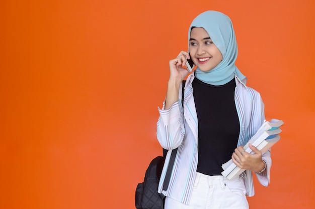 Asian muslim woman standing with backpack holding books while having a phone call