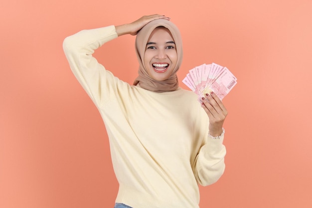 Asian muslim woman in sweater showing cash money in indonesian rupiah banknotes and holding head