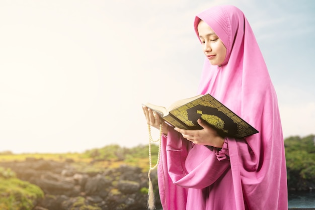 Asian Muslim woman in a veil holding prayer beads and reading the Quran at outdoor