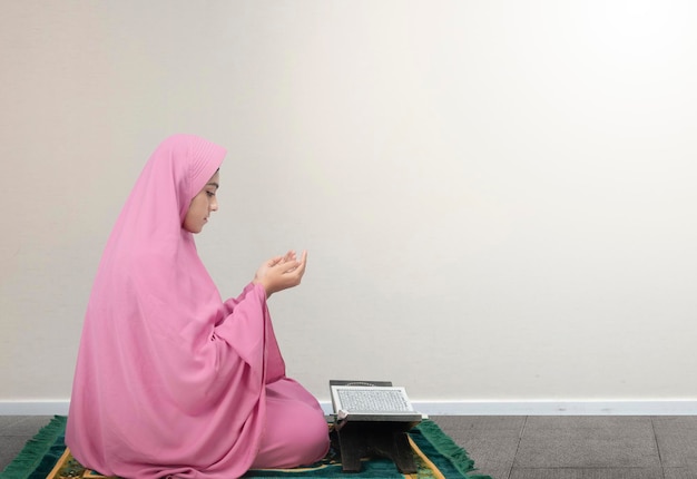 Asian Muslim woman in a veil sitting while raised hands and praying with white wall background