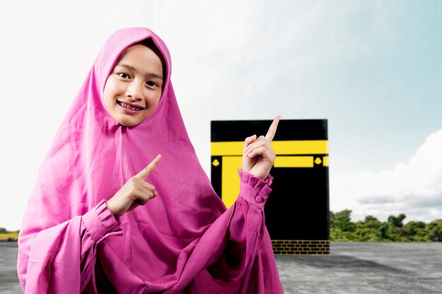 Asian muslim woman in a veil standing and pointing something\
with kaaba background