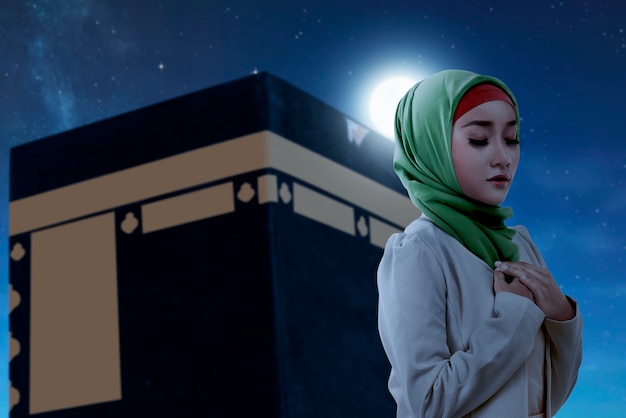 Donna musulmana asiatica con un velo in piedi e pregando con la vista della kaaba e lo sfondo della scena notturna