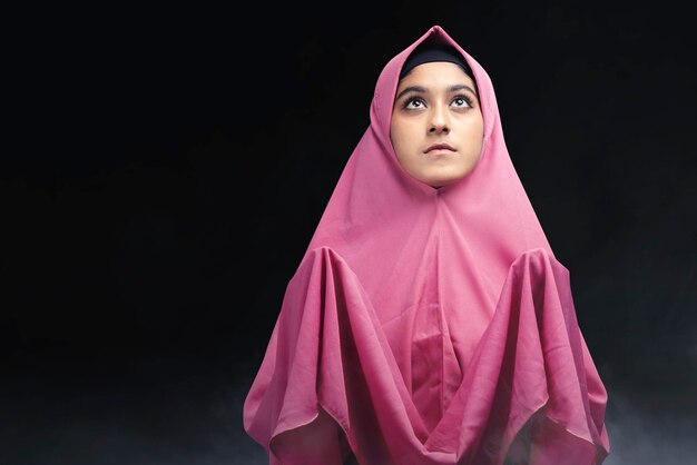 Asian muslim woman in a veil standing while raised hands and\
praying with dark background