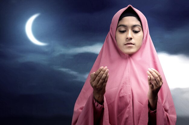 Asian muslim woman in a veil standing while raised hands and
praying with the night scene background