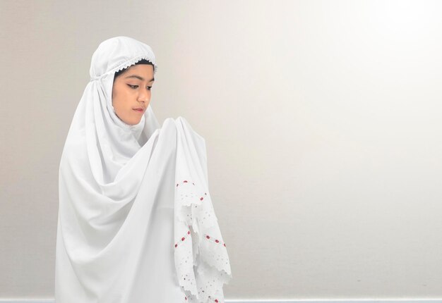 Asian muslim woman in a veil standing while raised hands and\
praying with white wall background