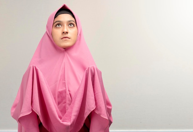 Asian Muslim woman in a veil standing while raised hands and praying with white wall background