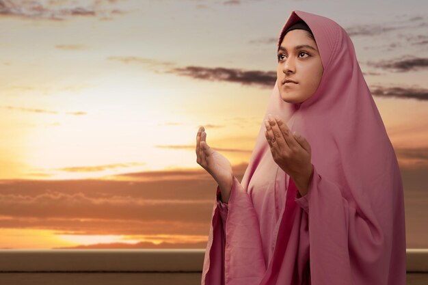 Asian muslim woman in a veil standing while raised hands and\
praying