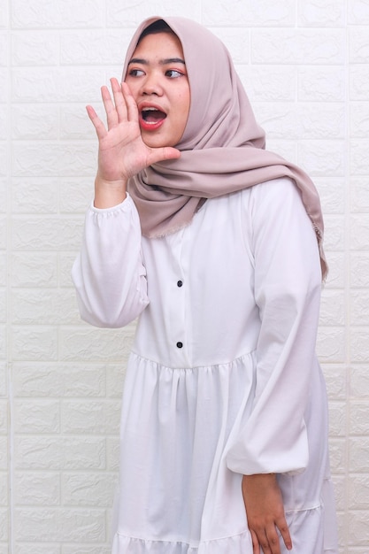 Asian Muslim woman wearing a headscarf shouting and screaming loud with a hand on her mouth