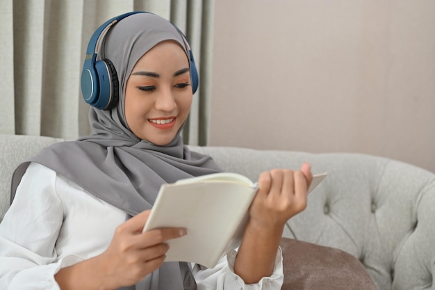 Asian Muslim woman wearing hijab and headphones reading a book in the living room