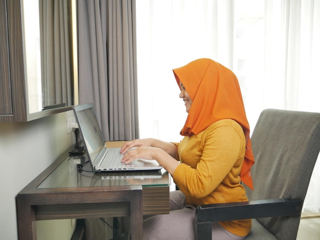 사진 히자브를 입은 아시아 무슬림 여성이 노트북에서 타이핑하는 동안 미소 짓고 집에서 일하는 개념