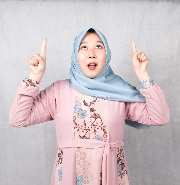 Asian Muslim woman wearing pink shirt and blue headscarf is promoting with both hands pointing up