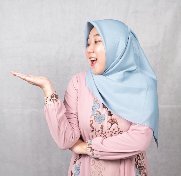 Asian Muslim woman wearing pink shirt is promoting with one hand pointing to the right in shock