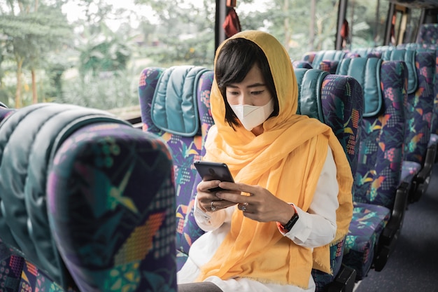 Азиатская мусульманка в маске использует свой мобильный телефон во время езды на автобусе