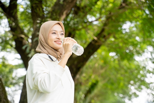 Donna musulmana asiatica con sciarpa per la testa che beve una bottiglia d'acqua durante l'allenamento all'aperto