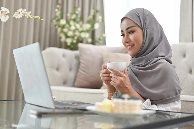 Азиатская мусульманка, работающая в гостиной, держа чашку кофе и используя портативный компьютер