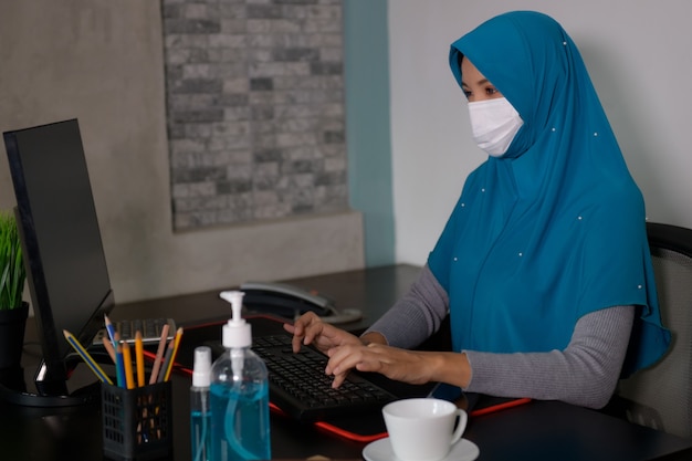 Азиатские мусульманки сидят и работают Дома во время распространения коронавируса (Covid-19) она носит маску и использует спирт для мытья рук. Для предотвращения микробов