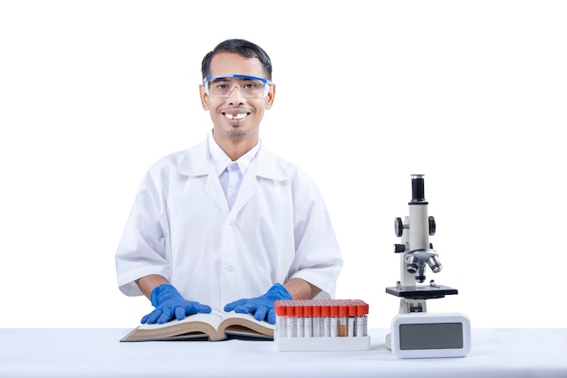 흰색 배경 위에 격리된 책상 위에 현미경과 의료 튜브 랙이 있는 책을 들고 서 있는 아시아 괴상한 과학자