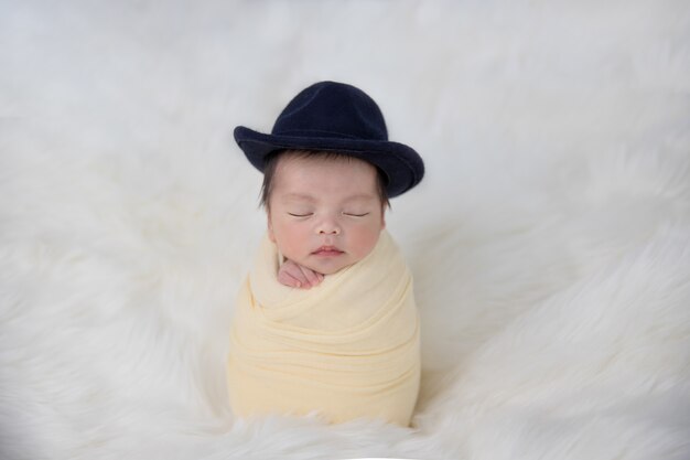 Asian Newborn Baby Wearing A Hat In Yellow Swaddle Sleep On White Fluffy Rug