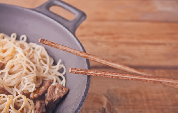 Asian noodles with beef, vegetables in wok with chopsticks, rustic wooden background. Asian style dinner. Chinese Japanese noodles