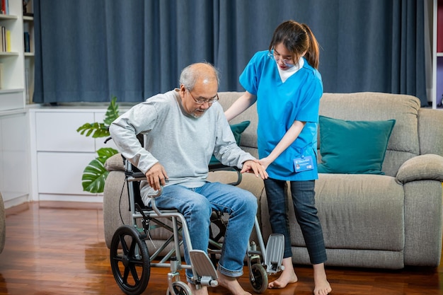 Photo asian nurse assisting helping senior man patient get up from wheelchair for practice walking at home, smiling doctor support old man to getting up, help handicapped elderly stand up, healthcare