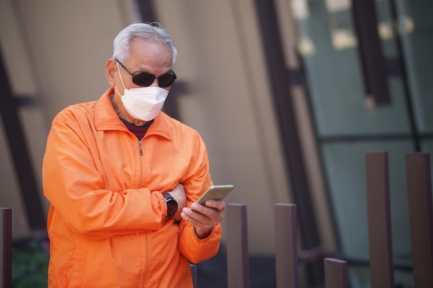 Фото Азиатский пожилой пожилой мужчина пожилой мужчина в маске с помощью мобильного смартфона мобильный телефон на открытом воздухе зрелый пенсионный образ жизни