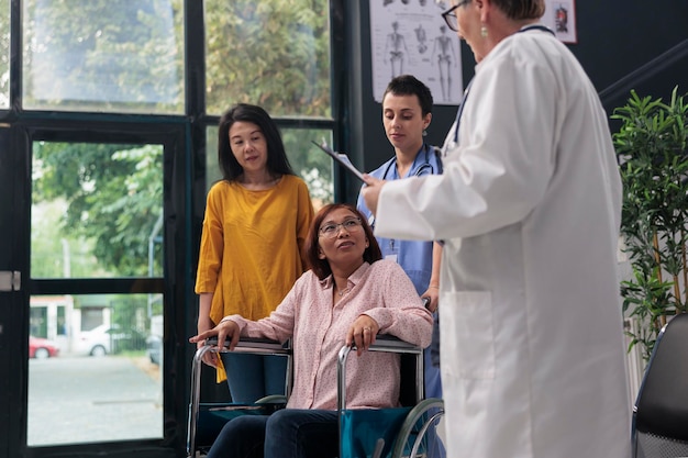 Asian old patient in wheelchair having medical consultation\
talking with elderly medic before physical examination in hospital\
waiting area. physician explaining disease diagnosis discussing\
treatment