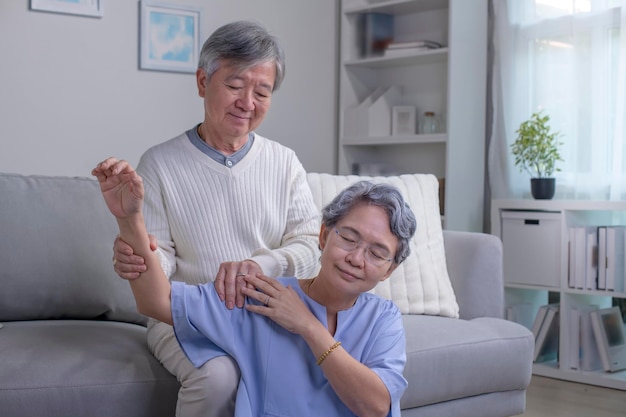 Asian old senior elderly wife sitting on floor having problem with suffer backache painful shoulder while care husband standing behind help massaging back