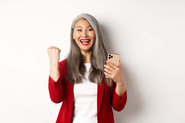 Asian old woman winning online, holding smartphone and making fist pump gesture to celebrate win, triumphing and smiling, standing over white background