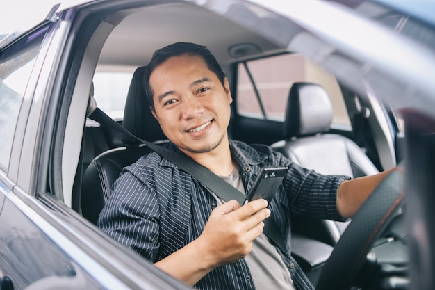 Asian online taxi driver looking at camera smiling and holding smart phone while sitting in car