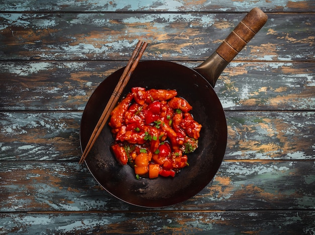 Фото Вкусный цыпленок в азиатском или китайском стиле в кисло-сладком соусе подается в черной деревенской сковороде вок на красочном деревянном фоне. концепция азиатского ужина, вид сверху, крупный план