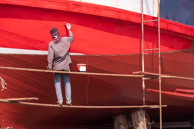 Asian painter on hanging platform is repainting hull of the old wooden fishing boat at shipyard