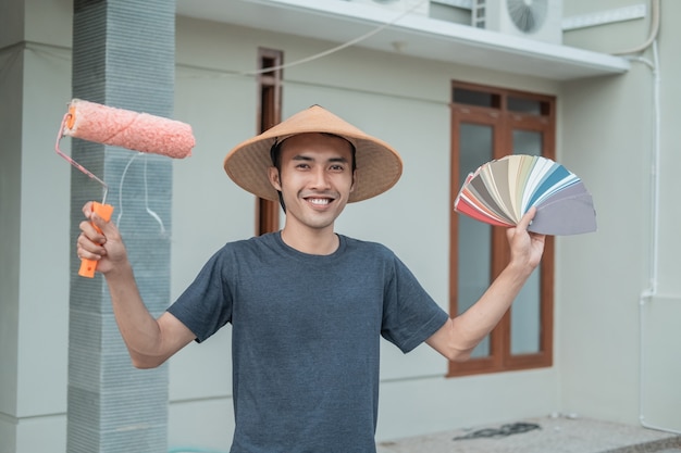 Asian painter smiles wearing a cap with Raise hands holding paint rollers and samples of paint colors