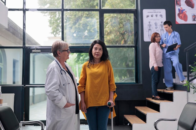 Photo asian patient trying to walk with walking cane in waiting area at health center, receiving help from senior doctor. old woman doing physiotherapy for physical injury rehabilitation. medicine support