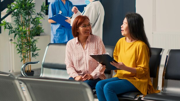 Asian Patients Talking About Medical Report Papers In Waiting Area, Preparing To Attend Consultation Appointment. Discussing About Healthcare Insurance And Treatment. Tripod Shot.