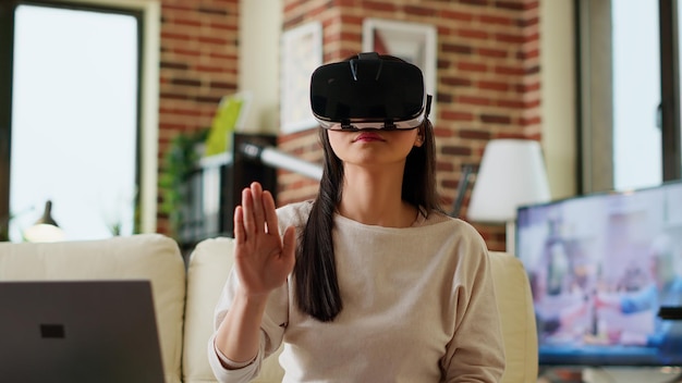 Asian person wearing futuristic VR goggles while interacting with cyber reality. Playful woman wearing virtual reality headset scrolling through metaverse while working from home. Handheld shot