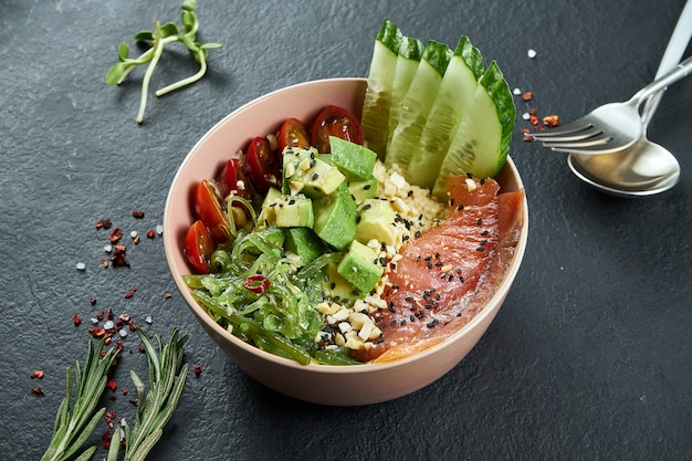 Ciotola di poke asiatica con salmone, avocado, pomodorini, cetrioli e wakame su una superficie nera