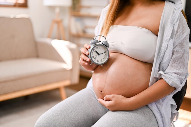 An Asian pregnant woman holding her belly and alarm clock awaiting baby baby's due date