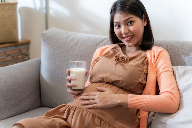 Una donna incinta asiatica che sorride e che si siede sul divano tiene un bicchiere di latte con sentimento felice e rilassato.