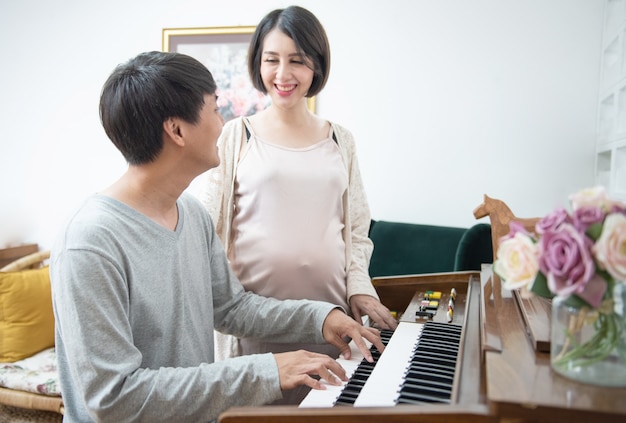 Asian pregnant woman stand side of man play piano at home