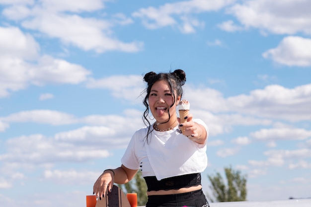 Ragazza graziosa asiatica che mangia il gelato nel parco, abbigliamento casual e skateboard
