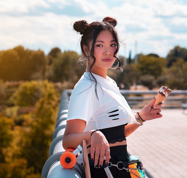 Ragazza graziosa asiatica che mangia il gelato nel parco, abbigliamento casual e skateboard