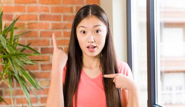 Asian pretty woman at home against brick wall with a plant