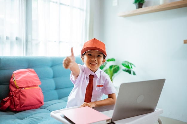 Studentessa asiatica delle elementari che mostra i pollici in su mentre usa il laptop durante la sessione di lezione online indossando la sua uniforme scolastica