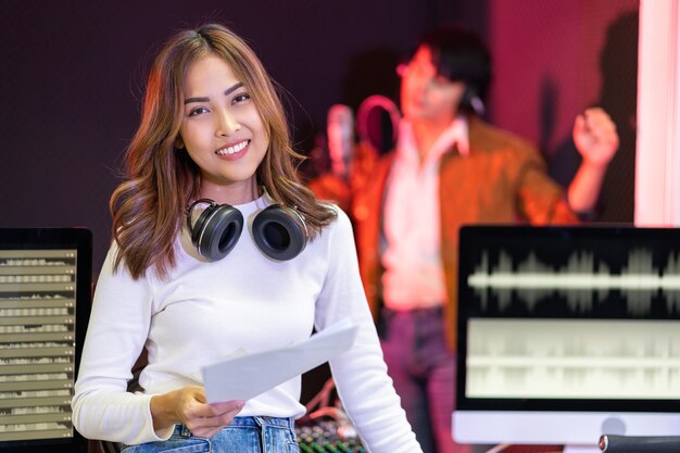 サウンドミキシングコンソールのそばに立っている白いシャツのアジアのプロデューサー女性幸せな女性の音楽作曲家アーティスト